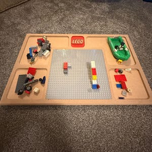 May include: A wooden LEGO tray with a gray baseplate and several compartments. The LEGO logo is in red. Various LEGO bricks and a green boat are arranged on the tray, ready for building.
