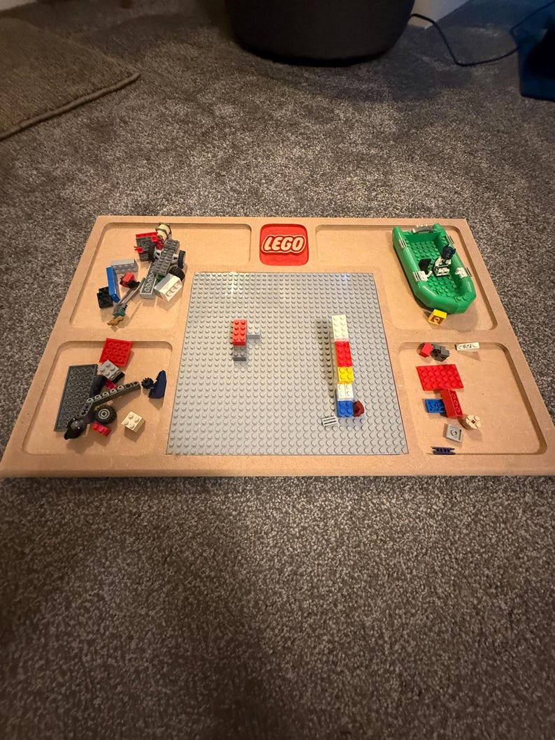 May include: A wooden LEGO tray with a gray baseplate and several compartments. The LEGO logo is in red. Various LEGO bricks and a green boat are arranged on the tray, ready for building.