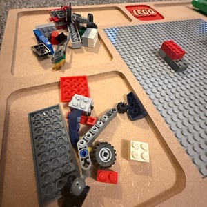 May include: A collection of colorful LEGO bricks and components scattered on a wooden tray. The tray has compartments and a built-in LEGO baseplate. The LEGO logo is visible. Various pieces include red, blue, gray, and white bricks, along with wheels and other structural elements.