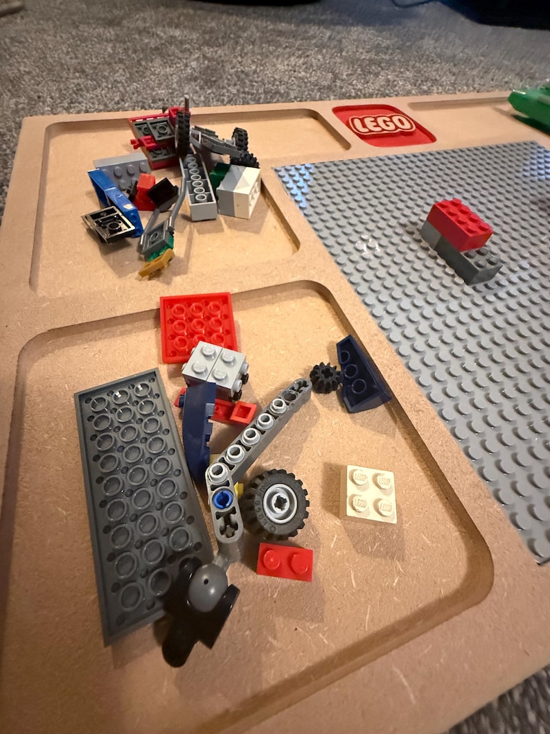 May include: A collection of colorful LEGO bricks and components scattered on a wooden tray. The tray has compartments and a built-in LEGO baseplate. The LEGO logo is visible. Various pieces include red, blue, gray, and white bricks, along with wheels and other structural elements.