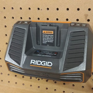 May include: A black and orange Ridgid 18V battery charger with a warning label and a power cord. The charger is mounted on a pegboard.