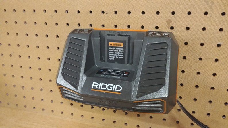 May include: A black and orange Ridgid 18V battery charger with a warning label and a power cord. The charger is mounted on a pegboard.