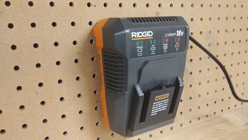 May include: A black and orange RIDGID 18v Lithium-Ion battery charger with a black power cord plugged in. The charger is mounted on a pegboard.