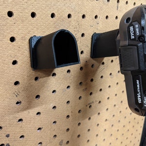 May include: Two black plastic wall mounts for holding power tools. The mount on the right is holding a black Ryobi 18V lithium battery with a 1.5Ah capacity. The battery has the text "RYOBI ONE+ 18V LITHIUM 1.5Ah" printed on it.