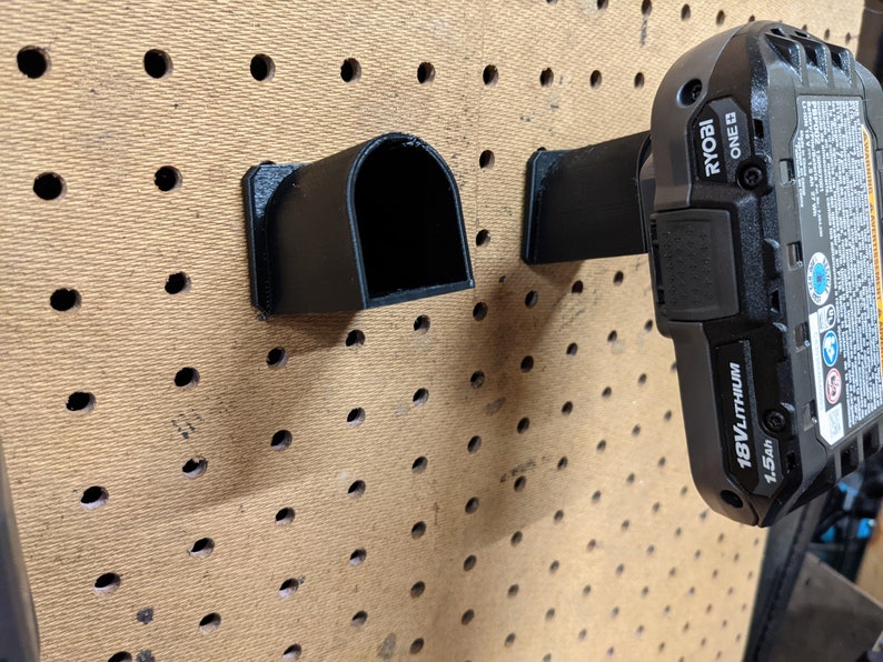 May include: Two black plastic wall mounts for holding power tools. The mount on the right is holding a black Ryobi 18V lithium battery with a 1.5Ah capacity. The battery has the text "RYOBI ONE+ 18V LITHIUM 1.5Ah" printed on it.
