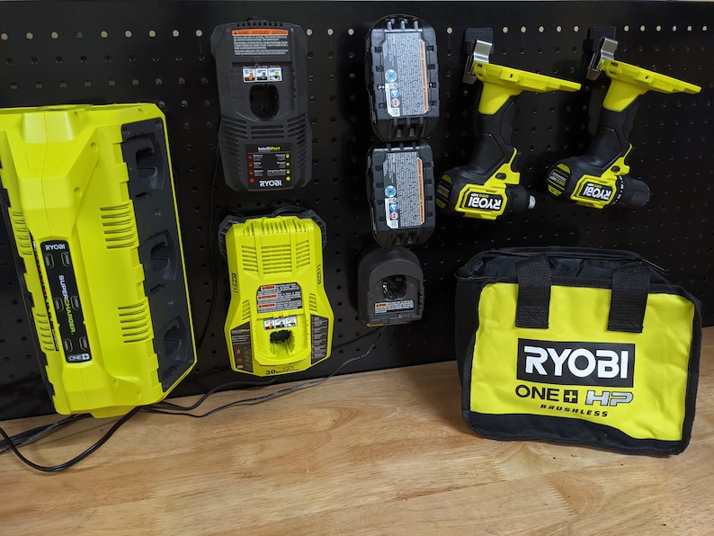 May include: A black pegboard with a variety of Ryobi tools, including a dual battery charger, a single battery charger, two batteries, two cordless drills, and a tool bag. The tools are all yellow and black and the bag has the Ryobi logo and the text "ONE+ HP Brushless".