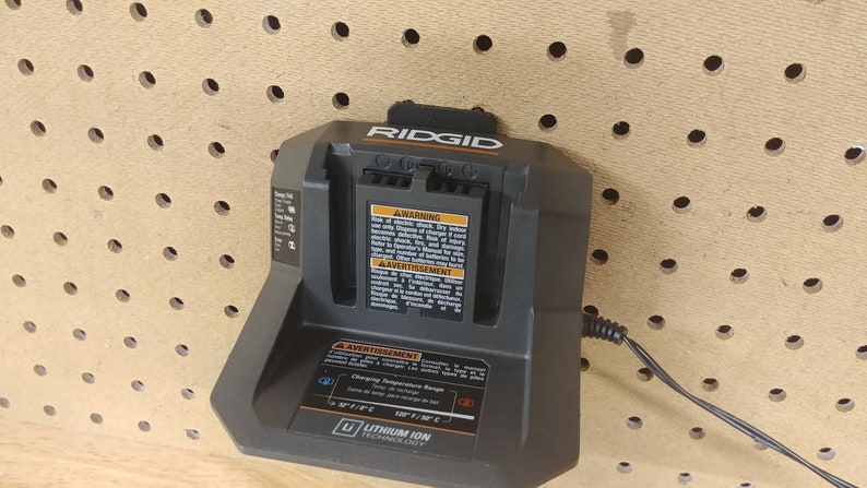 May include: A black Ridgid dual battery charger with a black power cord plugged in. The charger is mounted on a pegboard wall. The charger has a warning label and a lithium ion technology label.