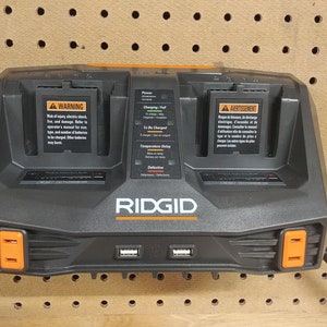 Puede incluir: Un cargador de bater&iacute;a de doble puerto RIDGID negro y naranja montado en una tabla perforada. El cargador tiene dos bah&iacute;as de carga, puertos USB e indicadores luminosos. El cargador tiene una etiqueta de advertencia y el logotipo de RIDGID.
