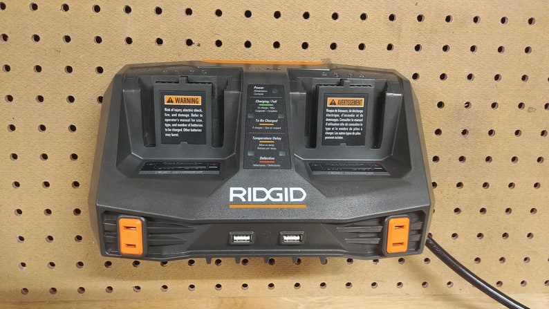 Puede incluir: Un cargador de bater&iacute;a de doble puerto RIDGID negro y naranja montado en una tabla perforada. El cargador tiene dos bah&iacute;as de carga, puertos USB e indicadores luminosos. El cargador tiene una etiqueta de advertencia y el logotipo de RIDGID.