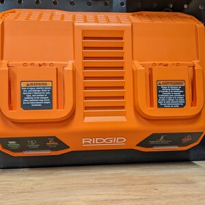 May include: An orange RIDGID dual-port battery charger. The charger has two charging ports, indicator lights, and warning labels. The charger is designed for power tool batteries.