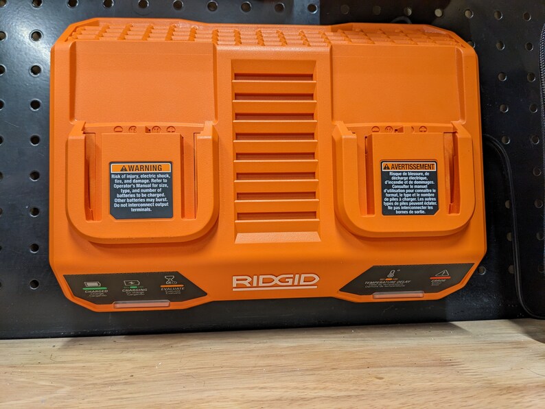 May include: An orange RIDGID dual-port battery charger. The charger has two charging ports, indicator lights, and warning labels. The charger is designed for power tool batteries.