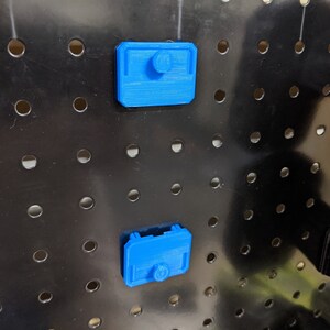 May include: Two blue plastic pegboard hooks with a rounded rectangular shape. The hooks are attached to a black pegboard.