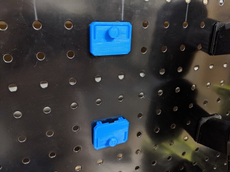 May include: Two blue plastic pegboard hooks with a rounded rectangular shape. The hooks are attached to a black pegboard.