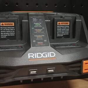 Puede incluir: Un cargador de bater&iacute;a RIDGID negro y naranja con dos compartimentos de carga, dos puertos USB y dos enchufes naranjas. El cargador tiene etiquetas de advertencia e indicadores de estado de carga. El logotipo de RIDGID es prominente.
