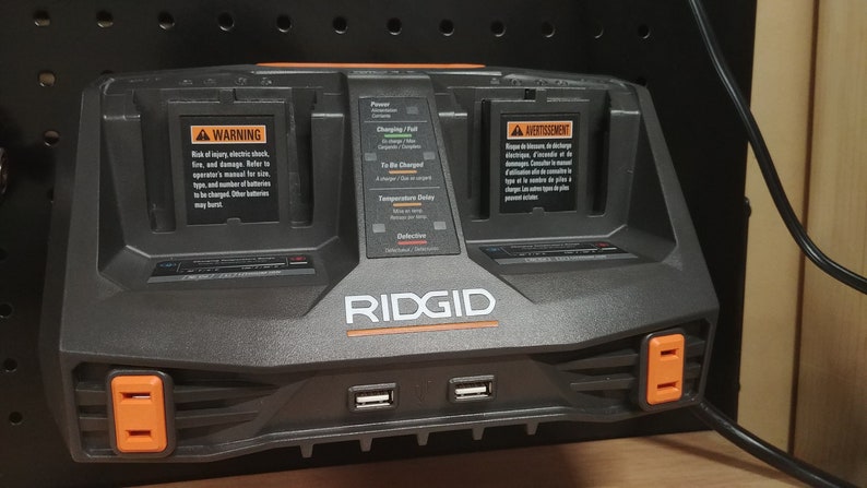 Puede incluir: Un cargador de bater&iacute;a RIDGID negro y naranja con dos compartimentos de carga, dos puertos USB y dos enchufes naranjas. El cargador tiene etiquetas de advertencia e indicadores de estado de carga. El logotipo de RIDGID es prominente.