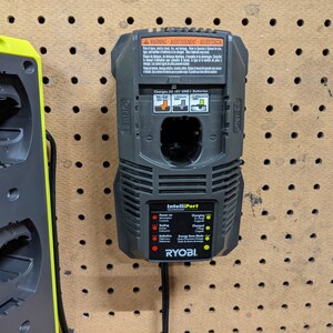 May include: A black Ryobi IntelliPort charger with a yellow indicator light. The charger is mounted on a pegboard wall and has a warning label with text in English, French, and Spanish.