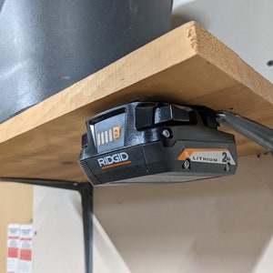 May include: A black and orange Ridgid 18V lithium-ion battery is mounted to a wooden shelf with a metal bracket. The battery has a charge indicator with four bars.