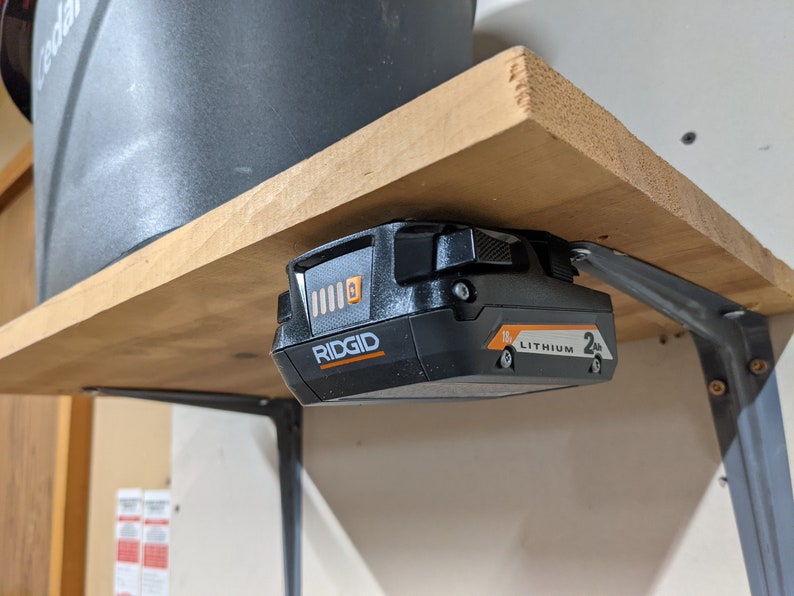 May include: A black and orange Ridgid 18V lithium-ion battery is mounted to a wooden shelf with a metal bracket. The battery has a charge indicator with four bars.