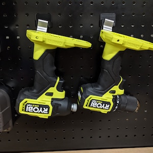 Può includere: Due trapani a batteria Ryobi ONE+ 18V gialli e neri appesi a una parete forata nera. Il trapano a sinistra è un avvitatore con una punta esagonale, e il trapano a destra è un trapano con un mandrino.