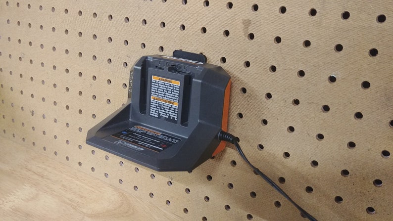 May include: A black and orange battery charger with a power cord plugged in. The charger is mounted on a pegboard wall.