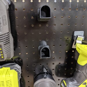 May include: A black pegboard with a variety of tools and accessories hanging on it. There are two black plastic tool holders, a black and yellow battery charger, a black and yellow battery, and a black and yellow drill. The pegboard is made of metal and has a black finish.