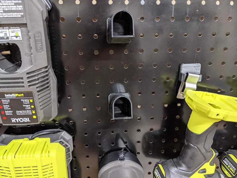 May include: A black pegboard with a variety of tools and accessories hanging on it. There are two black plastic tool holders, a black and yellow battery charger, a black and yellow battery, and a black and yellow drill. The pegboard is made of metal and has a black finish.