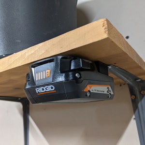 May include: A black and orange Ridgid 2.0Ah Lithium battery is mounted to a wooden shelf with a metal bracket.