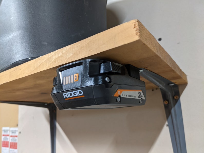 May include: A black and orange Ridgid 2.0Ah Lithium battery is mounted to a wooden shelf with a metal bracket.