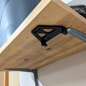 May include: A black 3D printed shelf bracket is attached to a wooden shelf with a silver metal bracket. The shelf is mounted to a wall.