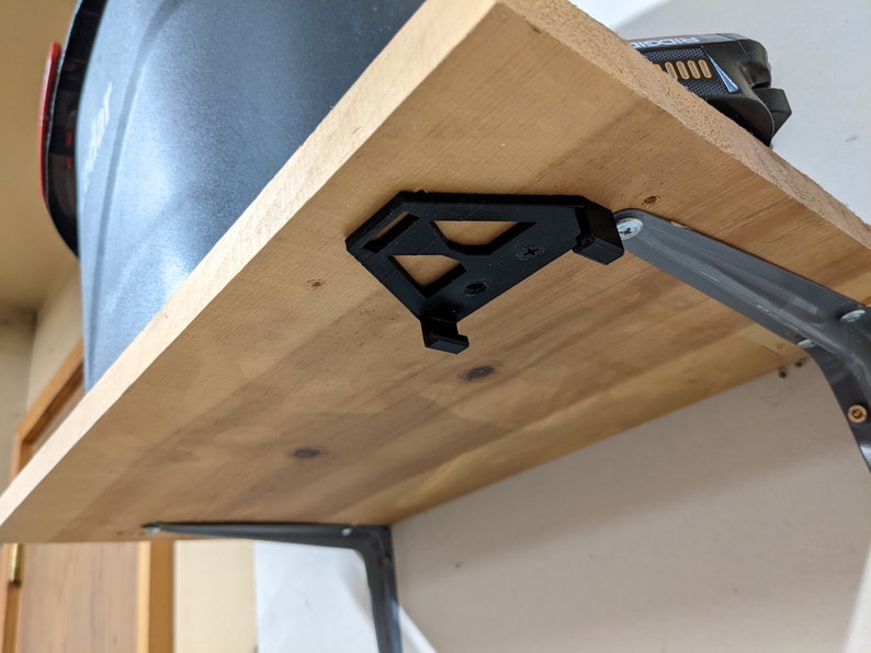 May include: A black 3D printed shelf bracket is attached to a wooden shelf with a silver metal bracket. The shelf is mounted to a wall.