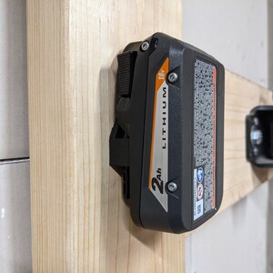 May include: A black and orange 2Ah 18V lithium battery is mounted on a wooden wall.