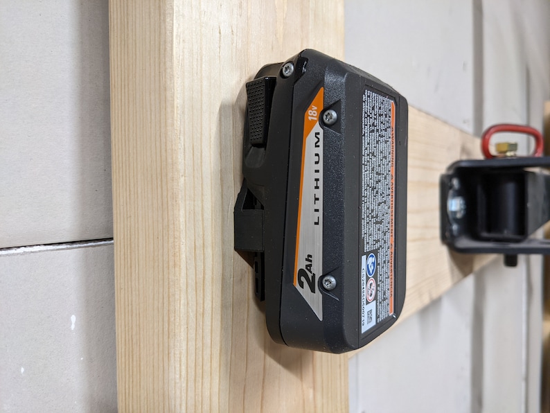 May include: A black and orange 2Ah 18V lithium battery is mounted on a wooden wall.