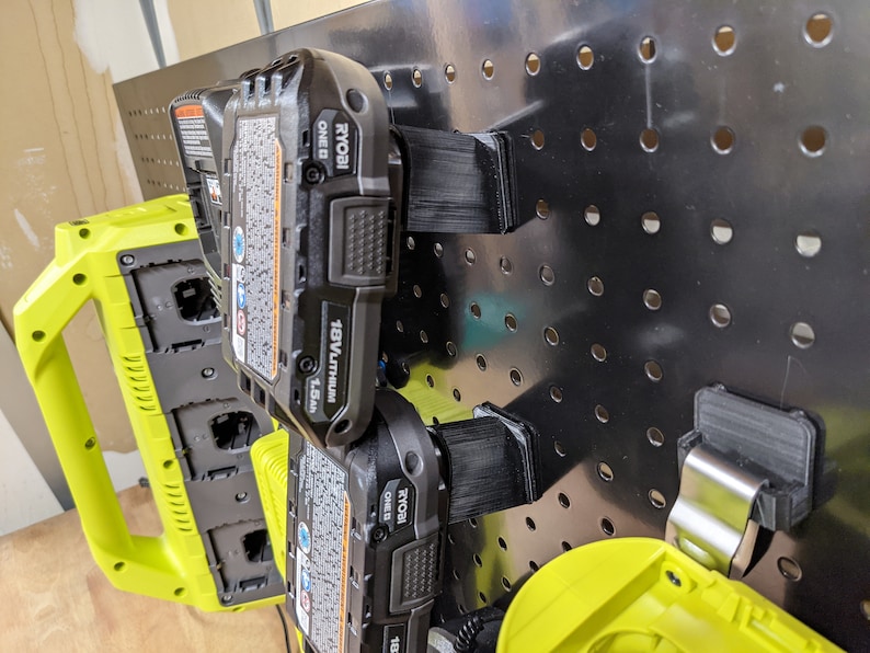 May include: Two black and yellow Ryobi 18V lithium-ion batteries are mounted on a black pegboard with black plastic battery holders. The batteries are labeled "RYOBI ONE+ 18V LITHIUM 1.5Ah".