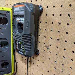 May include: Two Ryobi battery chargers, one black and one green, are mounted on a pegboard wall. The black charger has a warning label and the text "IntelliPort" and "ONE+" printed on it. The green charger has three battery slots.