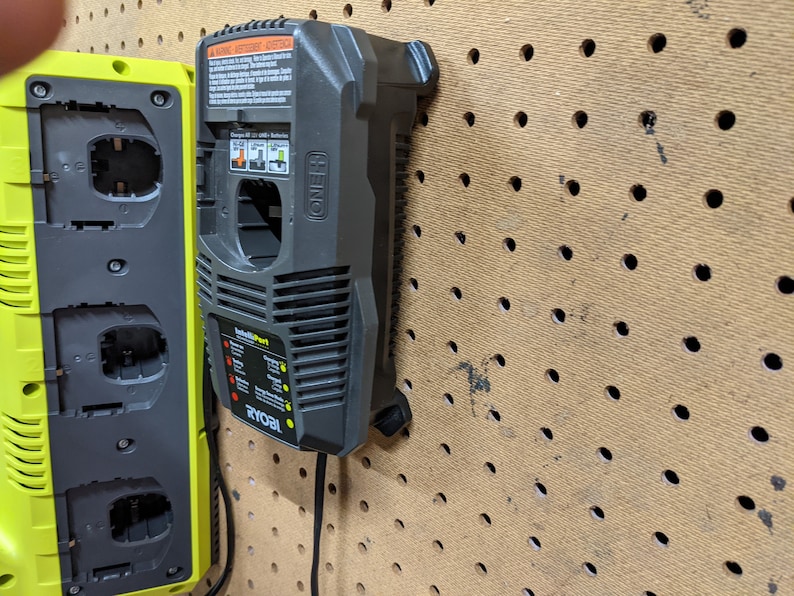 May include: Two Ryobi battery chargers, one black and one green, are mounted on a pegboard wall. The black charger has a warning label and the text "IntelliPort" and "ONE+" printed on it. The green charger has three battery slots.
