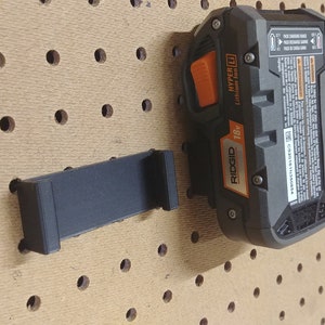 May include: A black 3D printed wall mount for a Ridgid 18V Hyper Lithium-Ion battery. The mount is attached to a pegboard.