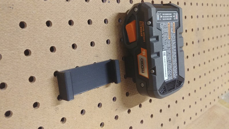 May include: A black 3D printed wall mount for a Ridgid 18V Hyper Lithium-Ion battery. The mount is attached to a pegboard.