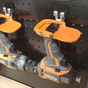 Op de afbeelding: Twee oranje en zwarte draadloze boormachines met bevestigde accupacks zijn gemonteerd op een zwart pegboard. De boormachines hebben de merknaam "RIDGID" erop gedrukt. Het pegboard heeft gelijkmatig verdeelde gaten.
