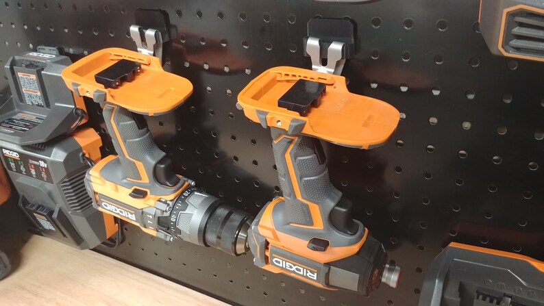 Op de afbeelding: Twee oranje en zwarte draadloze boormachines met bevestigde accupacks zijn gemonteerd op een zwart pegboard. De boormachines hebben de merknaam "RIDGID" erop gedrukt. Het pegboard heeft gelijkmatig verdeelde gaten.