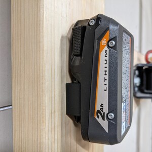 May include: A black and orange lithium-ion battery with a 2Ah capacity and 18V output. The battery is mounted on a wooden wall.