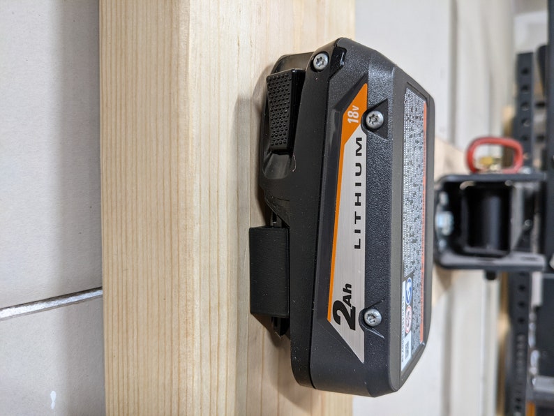 May include: A black and orange lithium-ion battery with a 2Ah capacity and 18V output. The battery is mounted on a wooden wall.