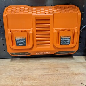 May include: Orange RIDGID dual-port battery charger. The charger has two charging ports with warning labels. The charger is mounted on a black pegboard. The charger has a power cord.