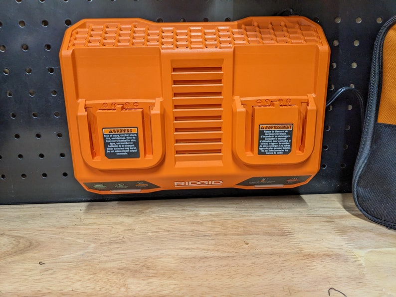 May include: Orange RIDGID dual-port battery charger. The charger has two charging ports with warning labels. The charger is mounted on a black pegboard. The charger has a power cord.