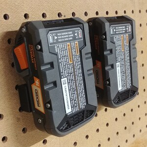 May include: Two black and orange Ridgid 18V Hyper Lithium-Ion batteries with a label that reads "Pack Charging Range 0-40&deg;C Pack Recharge Range 0-40&deg;C Pack de Carga Gama 0-40&deg;C" hanging on a pegboard.