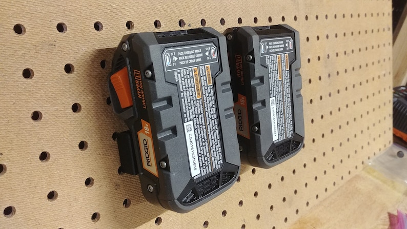 May include: Two black and orange Ridgid 18V Hyper Lithium-Ion batteries with a label that reads "Pack Charging Range 0-40&deg;C Pack Recharge Range 0-40&deg;C Pack de Carga Gama 0-40&deg;C" hanging on a pegboard.
