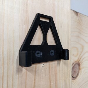 May include: A black plastic wall mount with two screws. The mount has a triangular shape with a cutout in the centre.