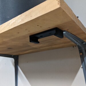 May include: A wooden shelf with a black metal bracket attached to the underside. The bracket has a small black plastic hook attached to it.