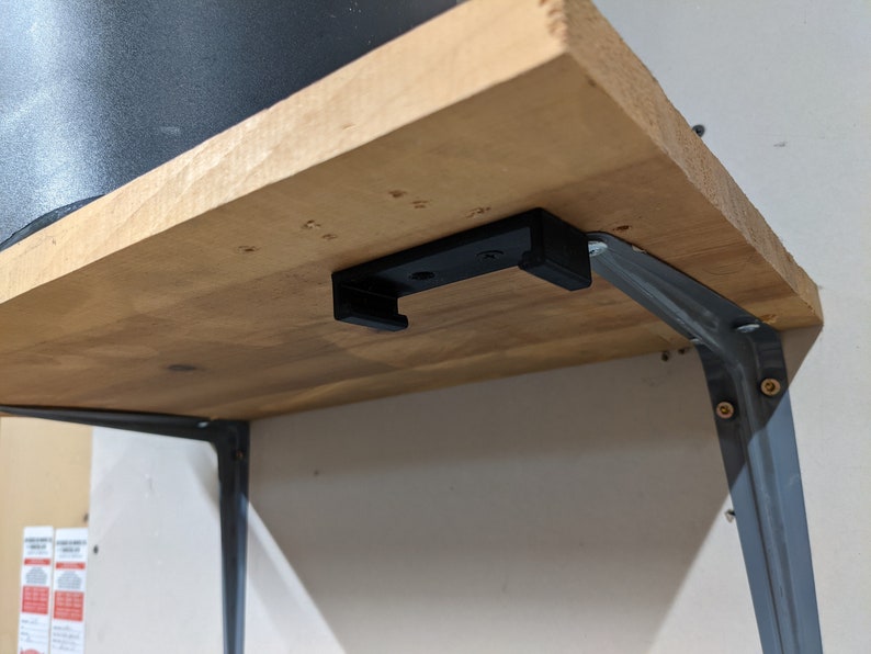 May include: A wooden shelf with a black metal bracket attached to the underside. The bracket has a small black plastic hook attached to it.