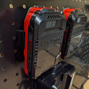 May include: Black and red Craftsman V20 Lithium Ion battery packs mounted on a black pegboard. The batteries are secured in black holders. The image shows two batteries in focus, with a third partially visible below.