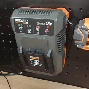 May include: A black and orange RIDGID 18V lithium-ion battery charger with a green light indicating charging. The charger is mounted on a black pegboard.